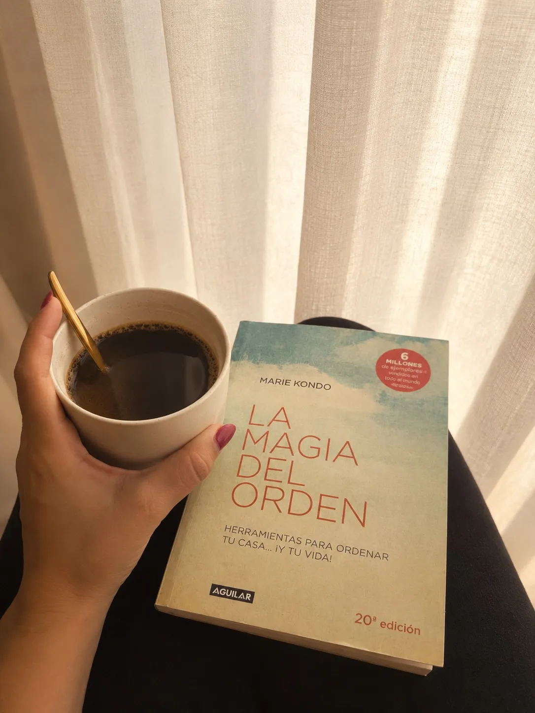 Mano de Paula sosteniendo un café junto a «La magia del orden» de Marie Kondo, a contraluz de una cortina de lino
