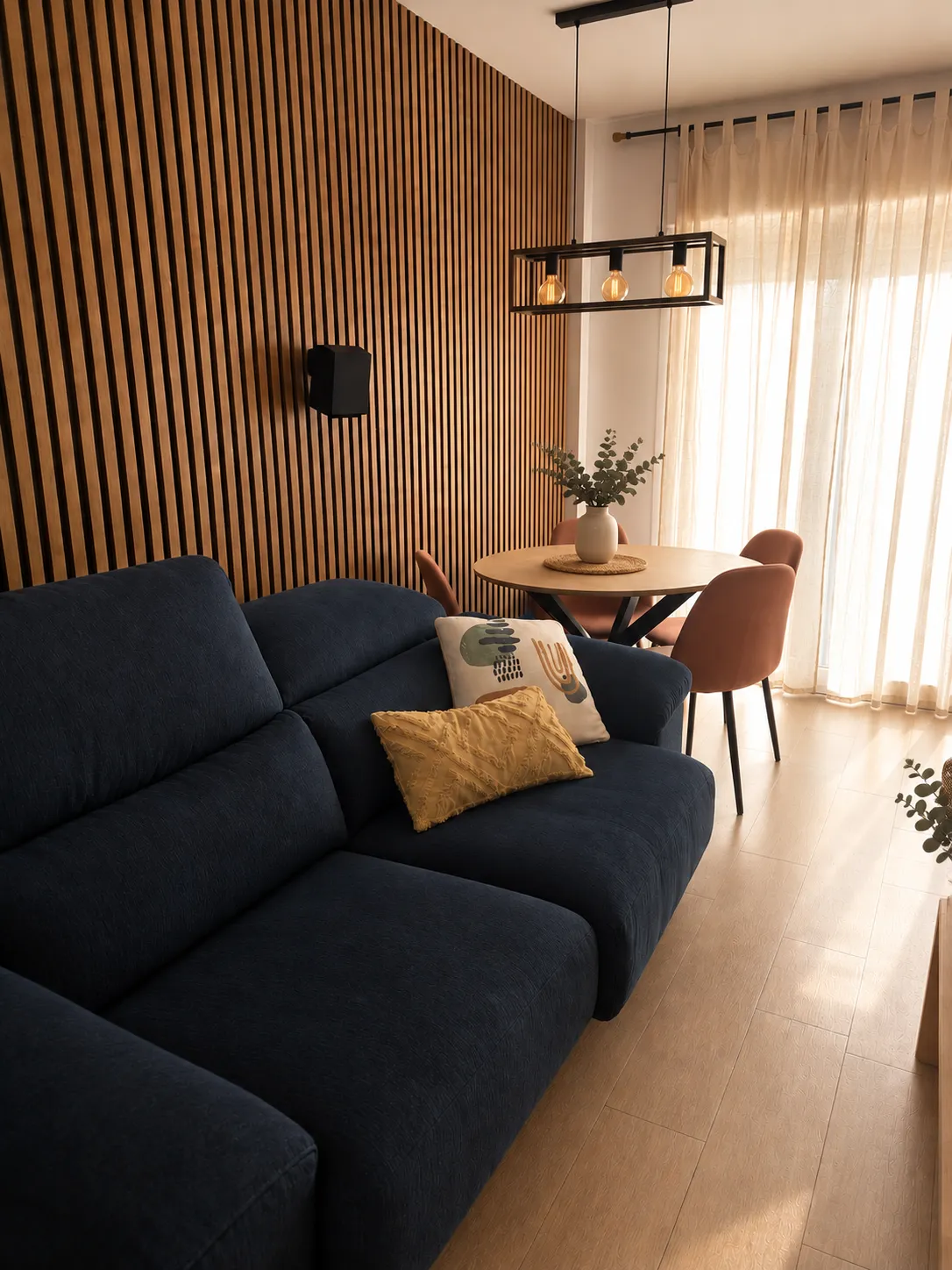 Salón-comedor de Casa Garpe: sofá azul noche junto a la pared de listones de madera, comedor con sillas terracota y cortinas de lino